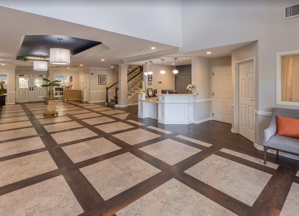 Spacious and well-lit senior living facility lobby with a checkered floor pattern, a white reception desk with pendant lights, a staircase in the background, a round table with a plant, and a gray chair with an orange cushion near the wall.
