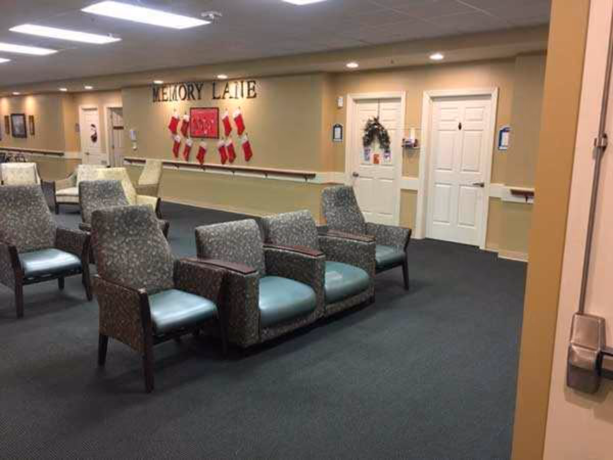 A seating area in a senior living facility with several cushioned chairs arranged in rows. The wall in the background has the words 'MEMORY LANE' displayed above a decoration of red stockings. There are two white doors with wreaths and informational signs on the walls. The carpet is dark, and the walls are painted beige.