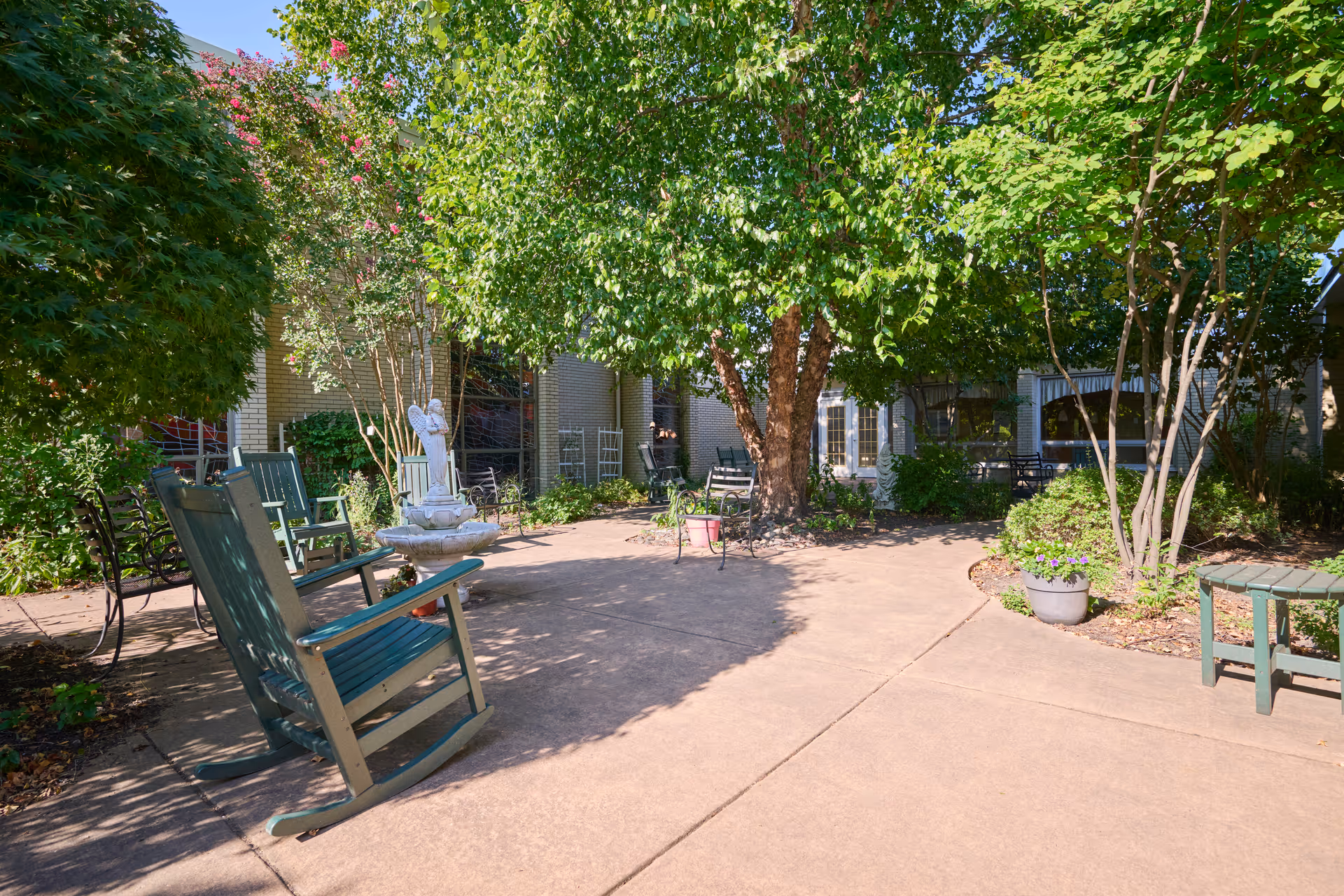 Sunlit courtyard with rocking chairs, potted plants, a small fountain, and trees outside the facility.