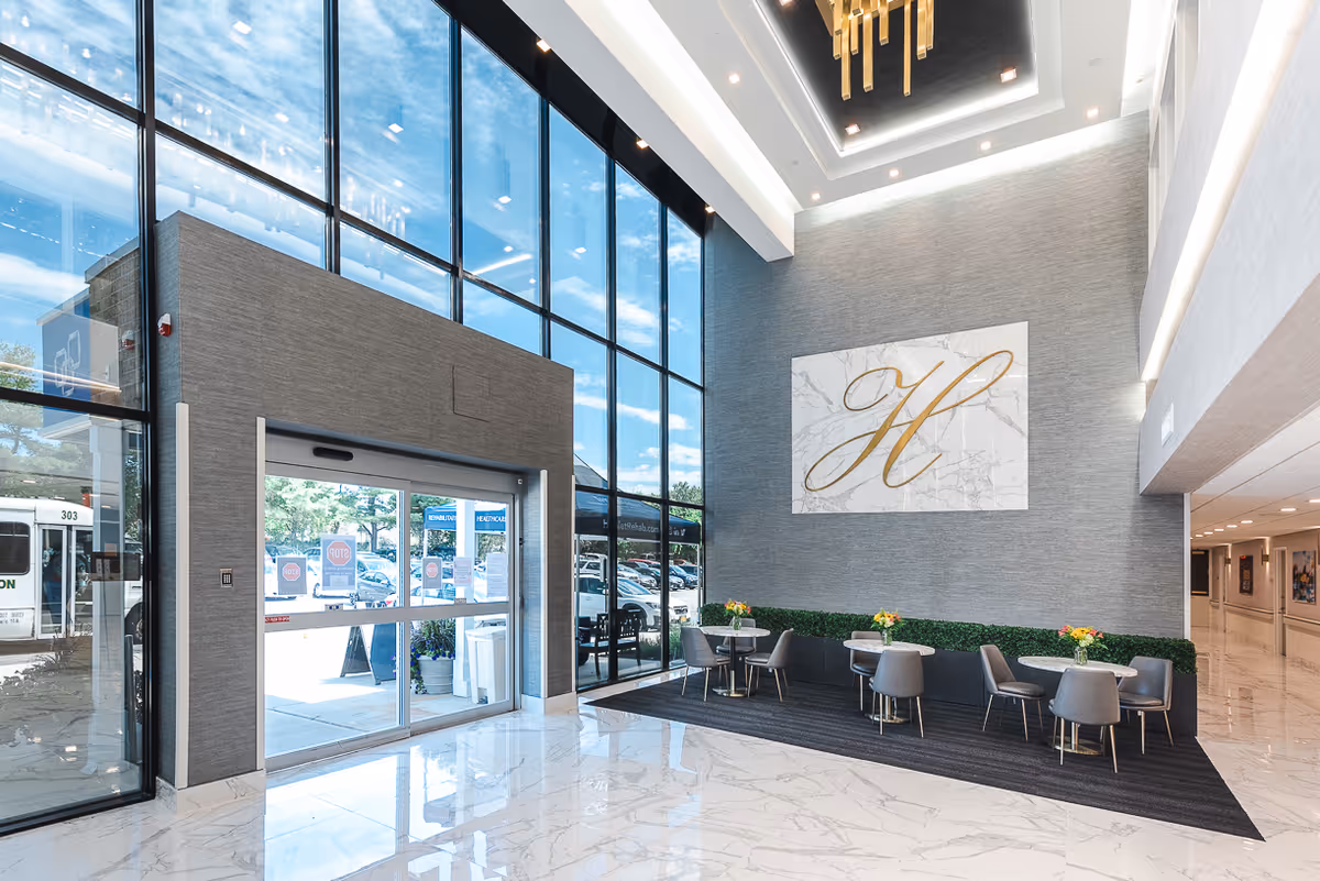 Bright and spacious lobby area with large floor-to-ceiling windows letting in natural light. The floor is covered with white marble tiles with gray veining. There is a seating area with small round tables and gray chairs, each table decorated with a small flower arrangement. On the wall above the seating area is a large decorative panel with a gold letter 'H' on a white marble background. The entrance features automatic sliding glass doors, and outside, a parking lot and a shuttle bus are visible.