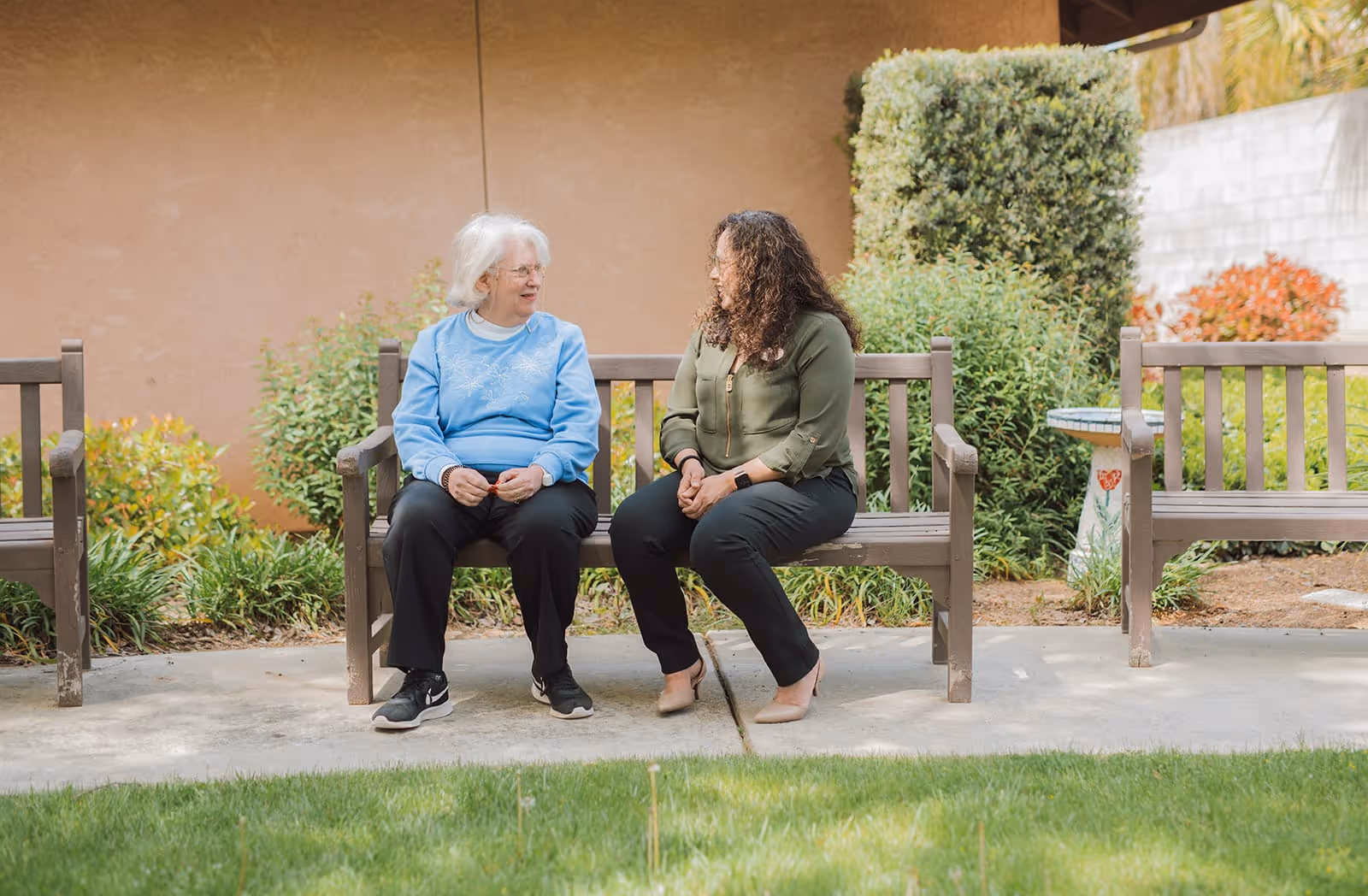 Two women sitting on a wooden bench outdoors, engaged in conversation. One woman has white hair and is wearing a blue sweater and black pants, while the other has curly hair and is wearing a green blouse and black pants. They are surrounded by greenery and plants with a beige wall in the background.