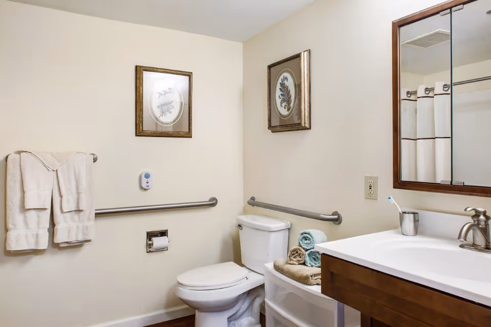 A clean bathroom with a toilet, a sink with a wooden cabinet, a mirror above the sink, two framed pictures on the walls, a towel rack with two white towels, a grab bar next to the toilet, and rolled towels placed on a small white storage unit.