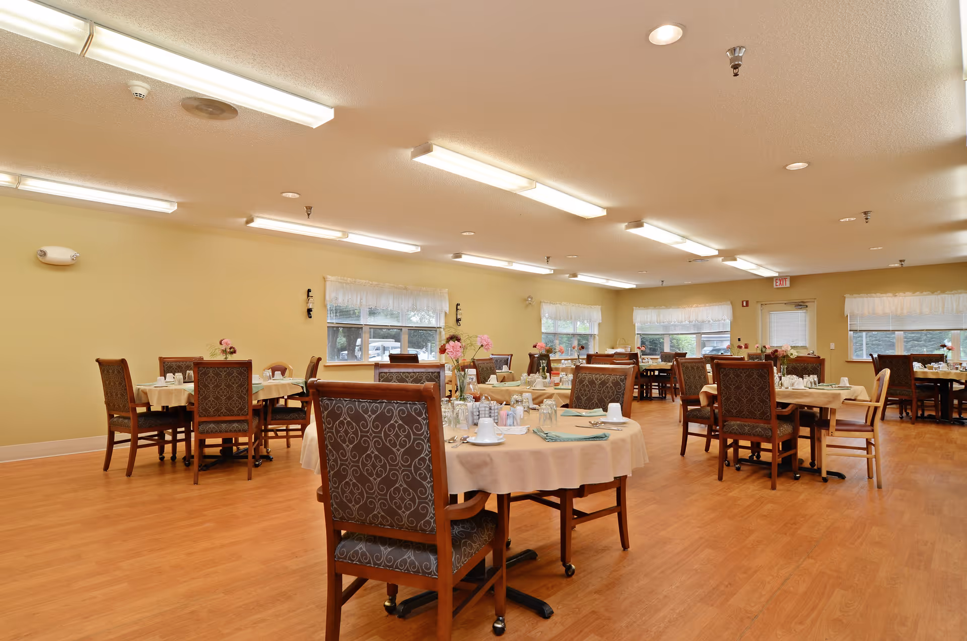 A spacious dining room with multiple round and rectangular tables covered with beige tablecloths. Each table is set with cups, glasses, and napkins, and decorated with small flower arrangements. The room has wooden flooring, beige walls, several windows with white curtains, and bright overhead fluorescent lighting.