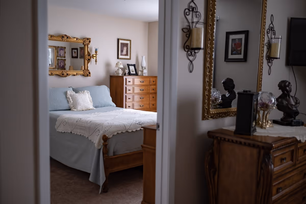 Bedroom with a made bed topped by a crocheted coverlet, wooden dressers, framed artwork and decorative mirrors.