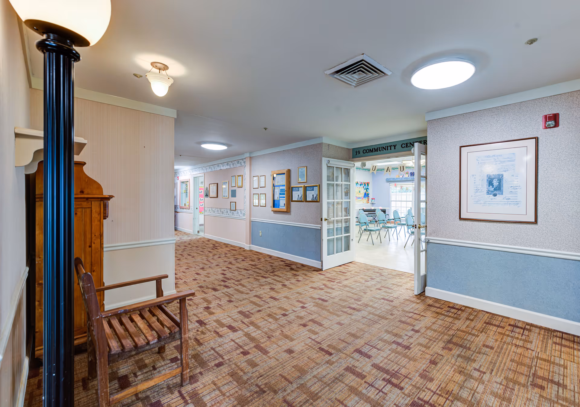 Carpeted hallway in a memory care facility leading to a community center with chairs and tables visible through open double doors.