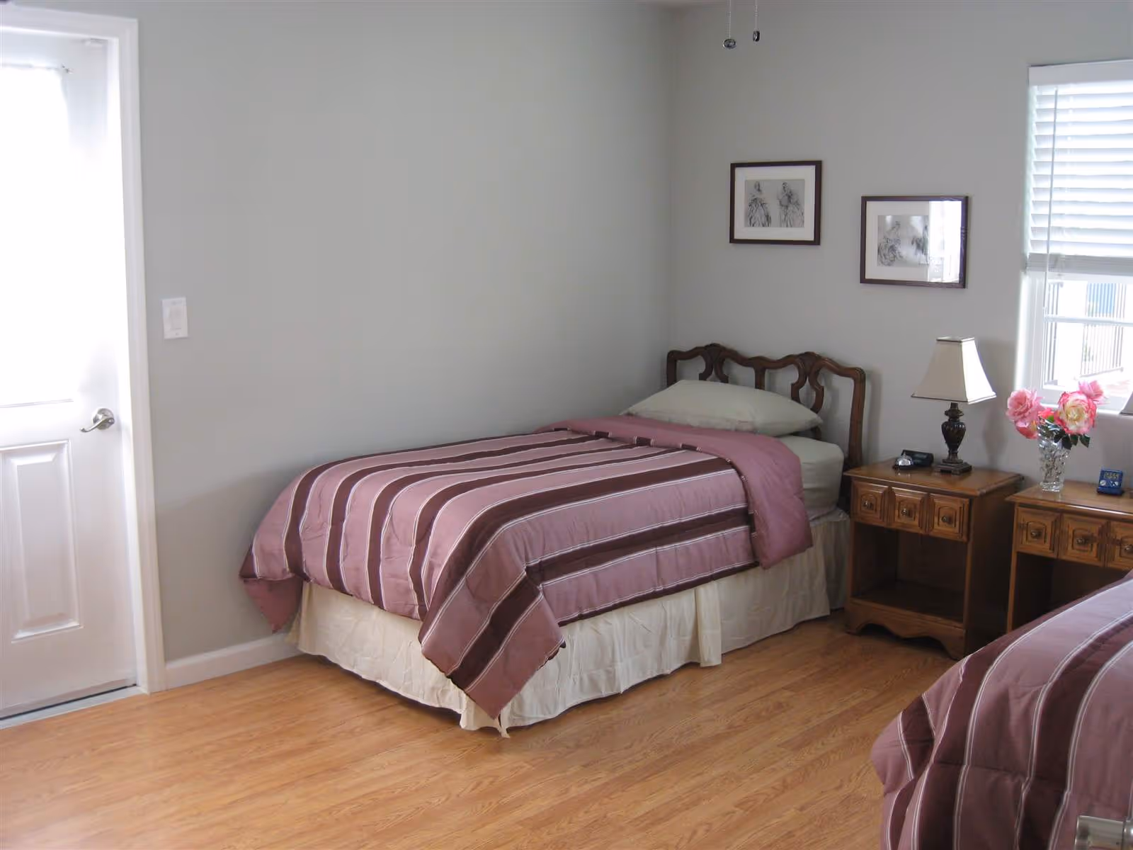 Bright bedroom with two twin beds covered in striped mauve comforters, wooden nightstands with a lamp and flowers, and framed art on the wall.