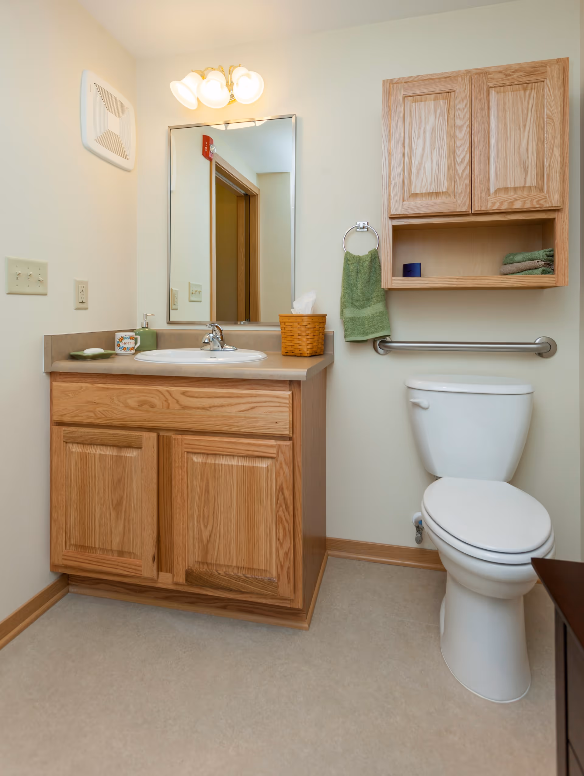 A clean bathroom featuring a wooden vanity with a sink, a mirror above it with three light fixtures, a toilet with a grab bar beside it, and a wooden wall cabinet with towels and a small container. A green hand towel hangs on a ring next to the cabinet.
