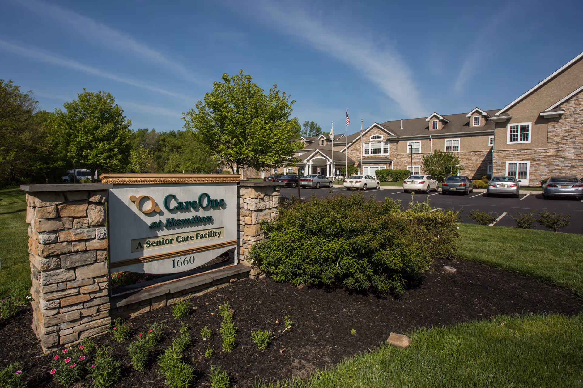 Exterior view of CareOne at Hamilton Assisted Living facility showing a stone sign with the facility name and address 1660, surrounded by landscaped bushes and flowers. The building is visible in the background with parked cars in the parking lot under a clear blue sky.