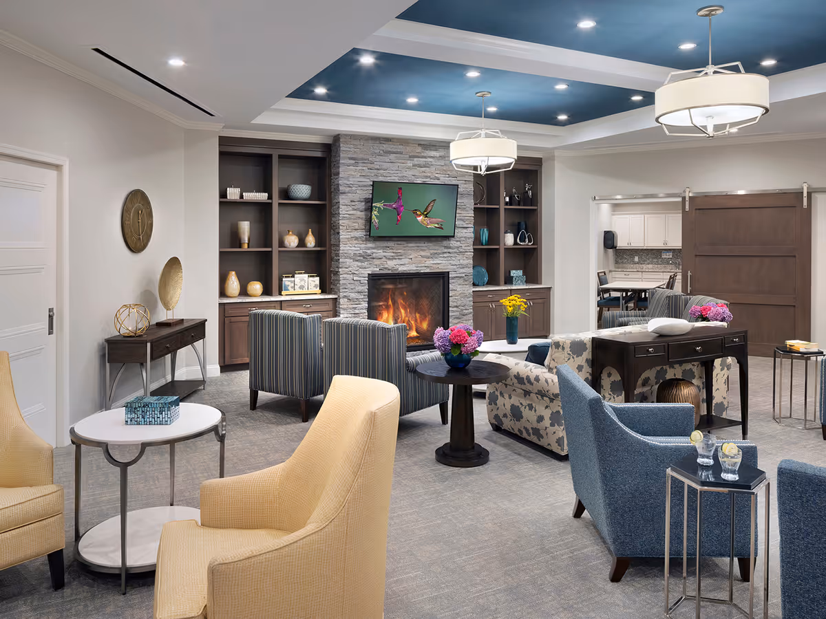 A cozy senior living facility common area with a stone fireplace, a wall-mounted TV displaying a hummingbird, and built-in shelves with decorative items. The room features various comfortable chairs and sofas arranged around small tables with flowers and drinks. The ceiling has recessed lighting and two hanging light fixtures, and a sliding wooden door leads to an adjacent room with a table and chairs.