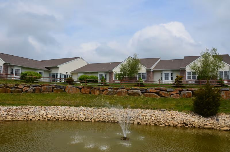 View of a single-story residential building with white siding and brown roofs behind a small pond with a water fountain. The building is surrounded by green grass, rocks, and a wooden fence, under a partly cloudy sky.