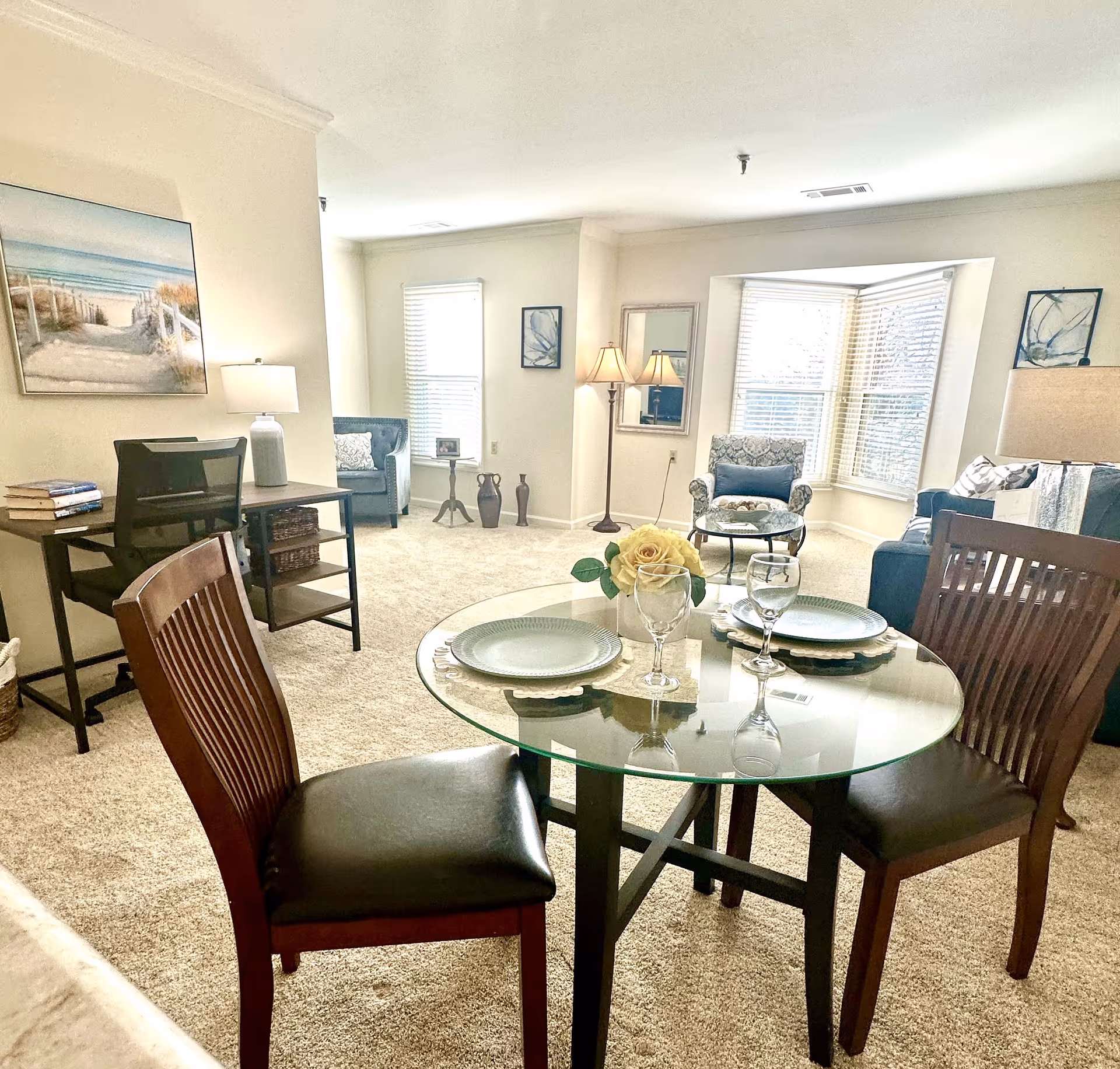 Bright open living area with a glass-top dining table set for two in the foreground and seating and a desk near bay windows in the background.
