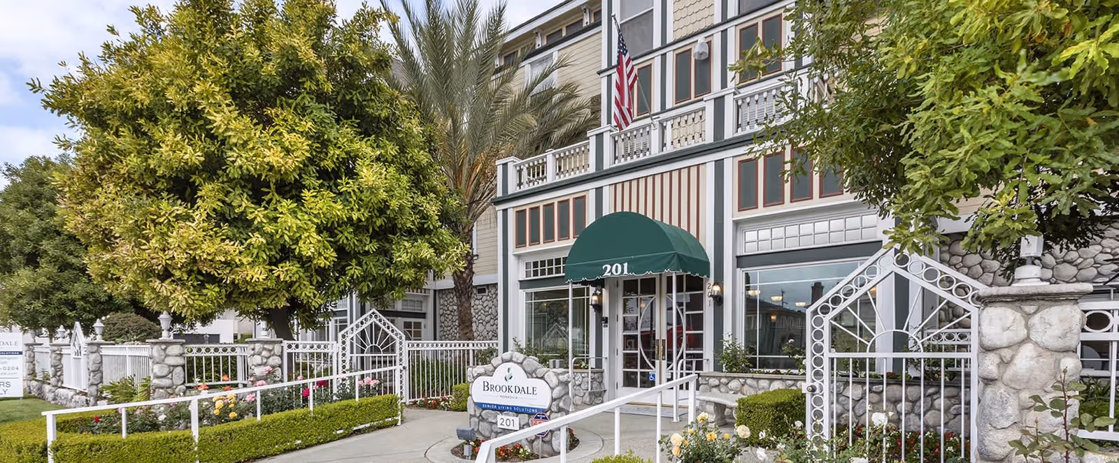 Exterior view of Brookdale Monrovia senior living facility showing the entrance with a green awning labeled 201, surrounded by stone pillars, white metal fencing, greenery, and an American flag above the entrance.