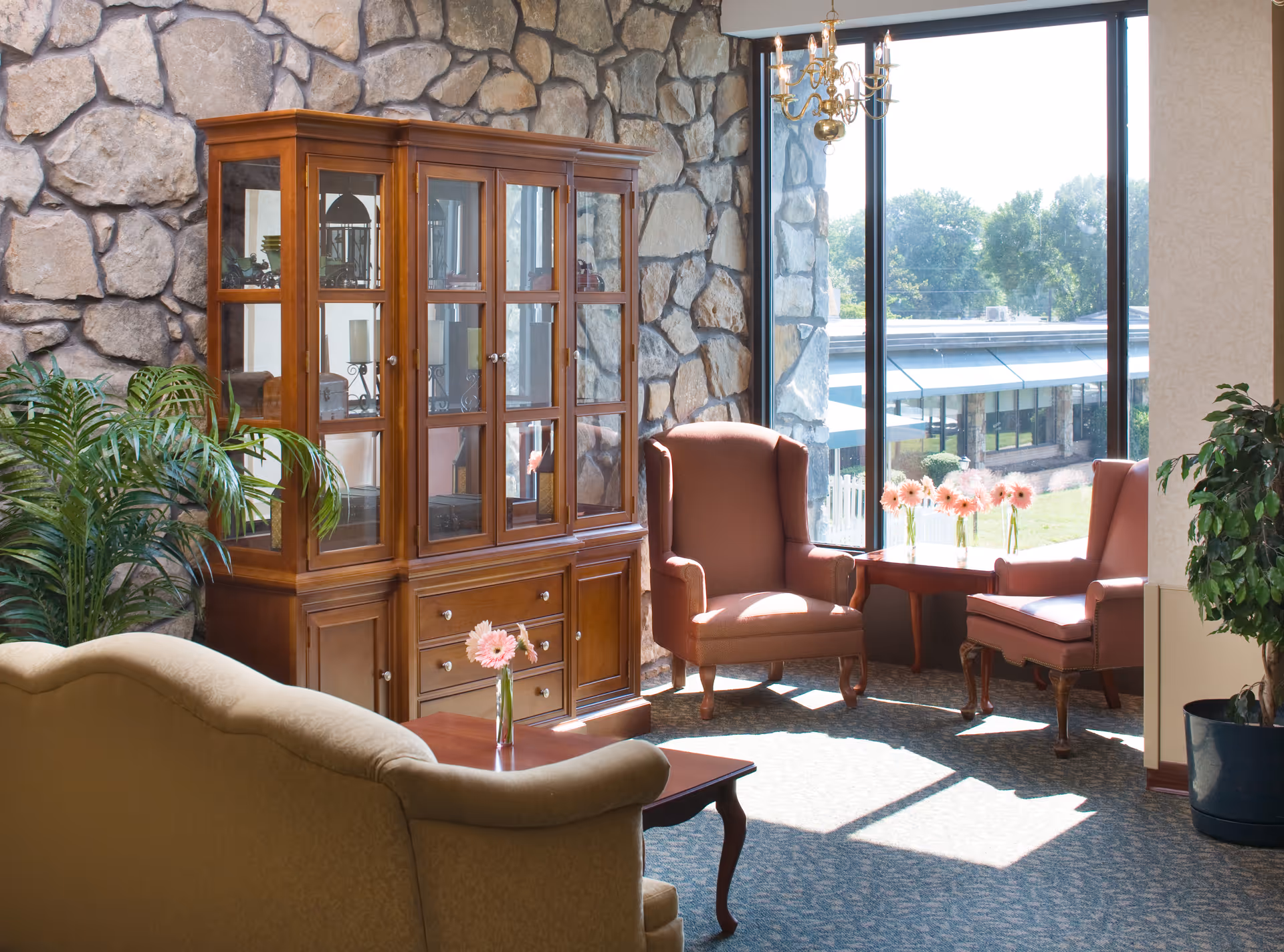A cozy sitting area in a senior living facility with a stone accent wall, a wooden glass-front cabinet, two upholstered armchairs, a small wooden table with pink flowers in vases, a beige sofa, and large windows letting in natural light with a view of greenery outside.