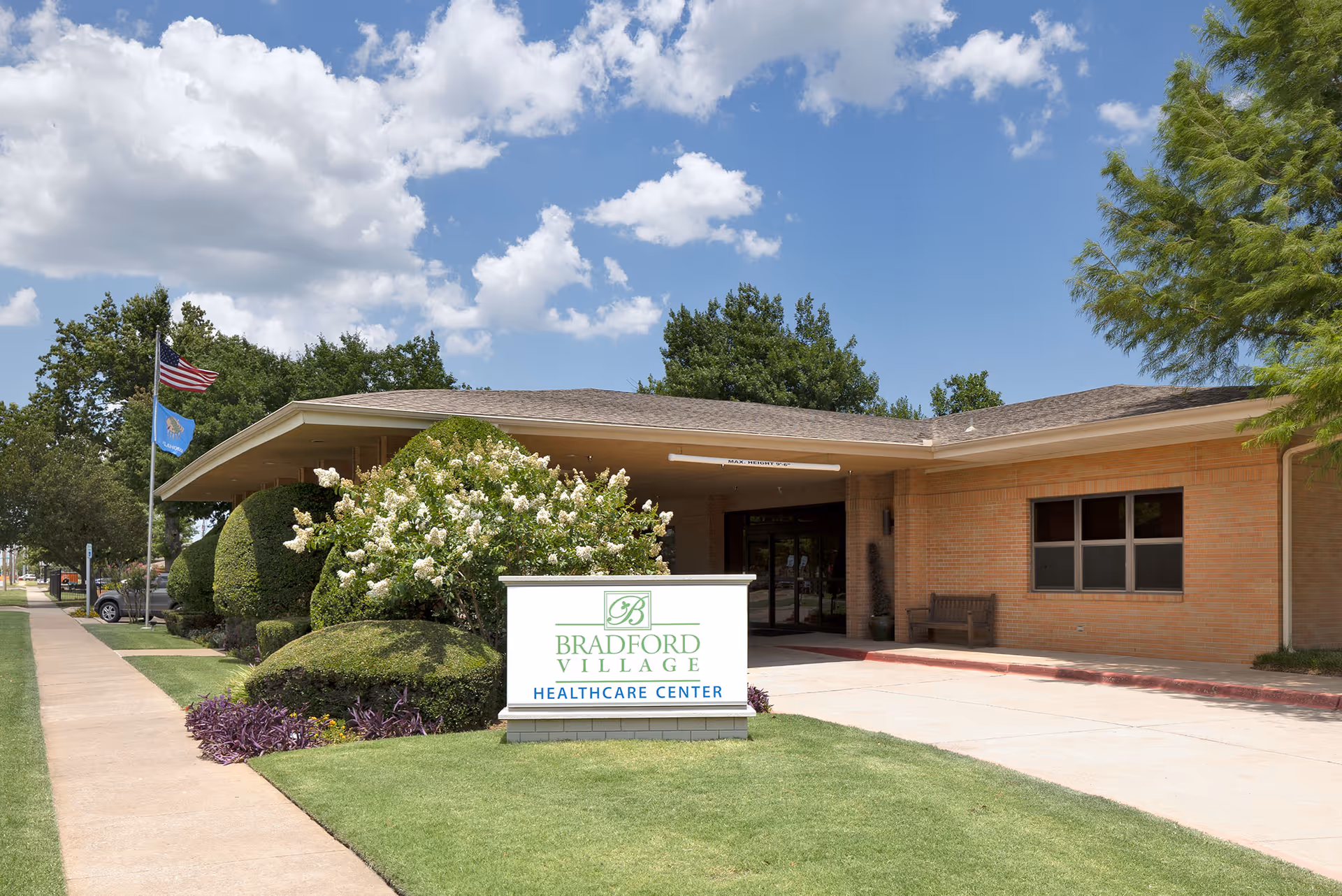 Exterior view of Bradford Village Healthcare Center building with a sign in front, well-maintained bushes and trees, a sidewalk, and two flagpoles with the American flag and another flag under a partly cloudy sky.