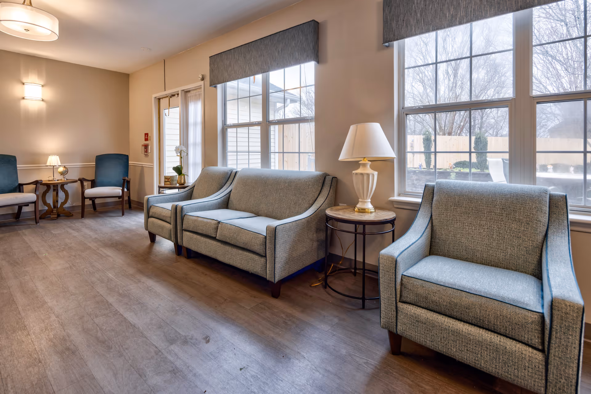A bright and cozy sitting area with light blue upholstered chairs and sofas arranged along the walls. There are two small round side tables, one with a white lamp and the other with a small plant and decorative items. Large windows let in natural light and show a fenced outdoor area with trees. The floor is wood, and the walls are painted a soft beige color.