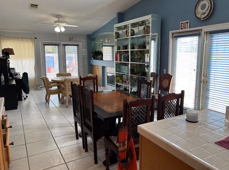 Interior view of a senior living facility dining and common area with a wooden dining table and chairs in the foreground, a glass display cabinet with plants and decorative items, a fireplace, and a round table with chairs near glass doors leading outside. The walls are painted blue, and there are ceiling fans and clocks visible.