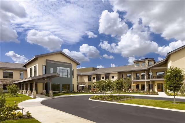 Front exterior of a two-story senior living building with a covered entrance, driveway, and landscaped grounds under a partly cloudy sky.