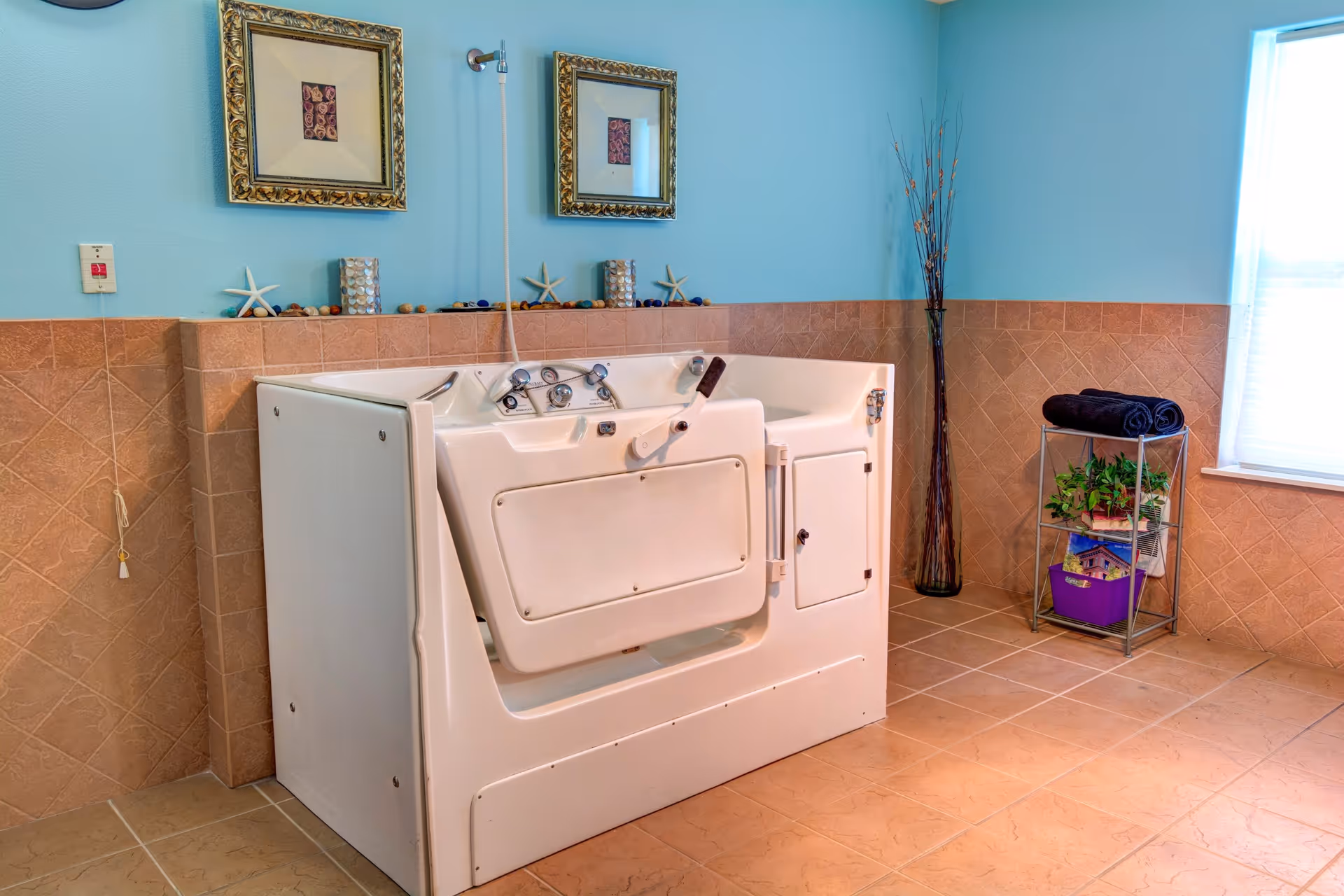 A walk-in bathtub in a bathroom with light blue walls and beige tiled floors and walls. The bathtub has a door with a handle for easy access. Above the tub are two framed pictures and decorative items including starfish and small stones. To the right, there is a metal shelf with rolled black towels, plants, and books. A tall vase with decorative branches stands in the corner near a window with blinds.