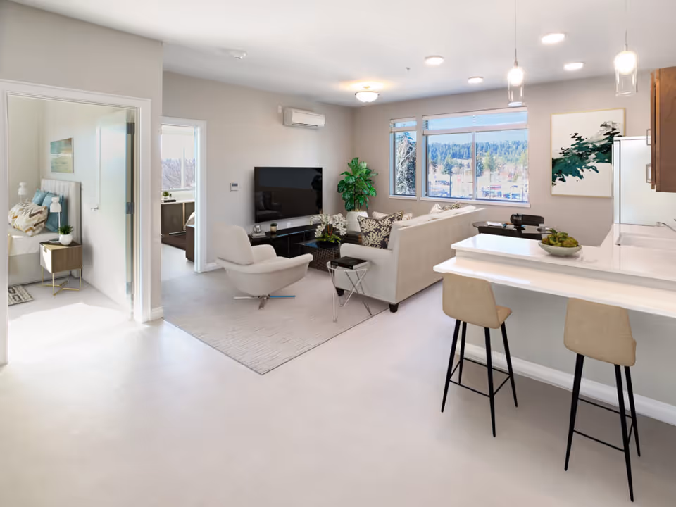Bright and modern open-concept living space in a senior living facility featuring a living room with a sofa, armchair, coffee table, and TV mounted on the wall. Adjacent to the living room is a kitchen area with a breakfast bar and two stools. Large windows provide natural light and a view of trees outside. A bedroom is visible through an open door on the left side of the image.
