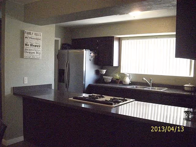 Kitchen with a central island cooktop, stainless steel refrigerator, sink beneath a long window and dark cabinetry.