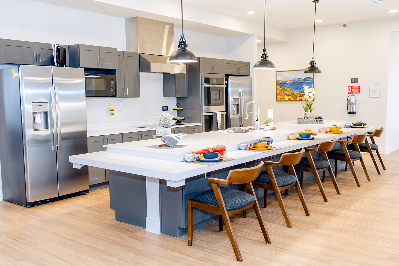 Modern kitchen with gray cabinets, stainless steel appliances including two refrigerators and a microwave, a long white island with a sink, and wooden chairs with blue cushions arranged along the island. The kitchen is well-lit with three pendant lights hanging above the island and a painting on the wall in the background.
