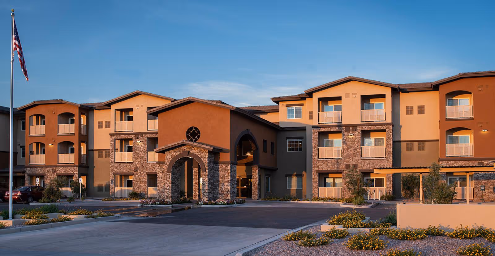 Exterior front of a three-story tan and brown senior living building with stone arch entrance, balconies, landscaped driveway, and an American flag.