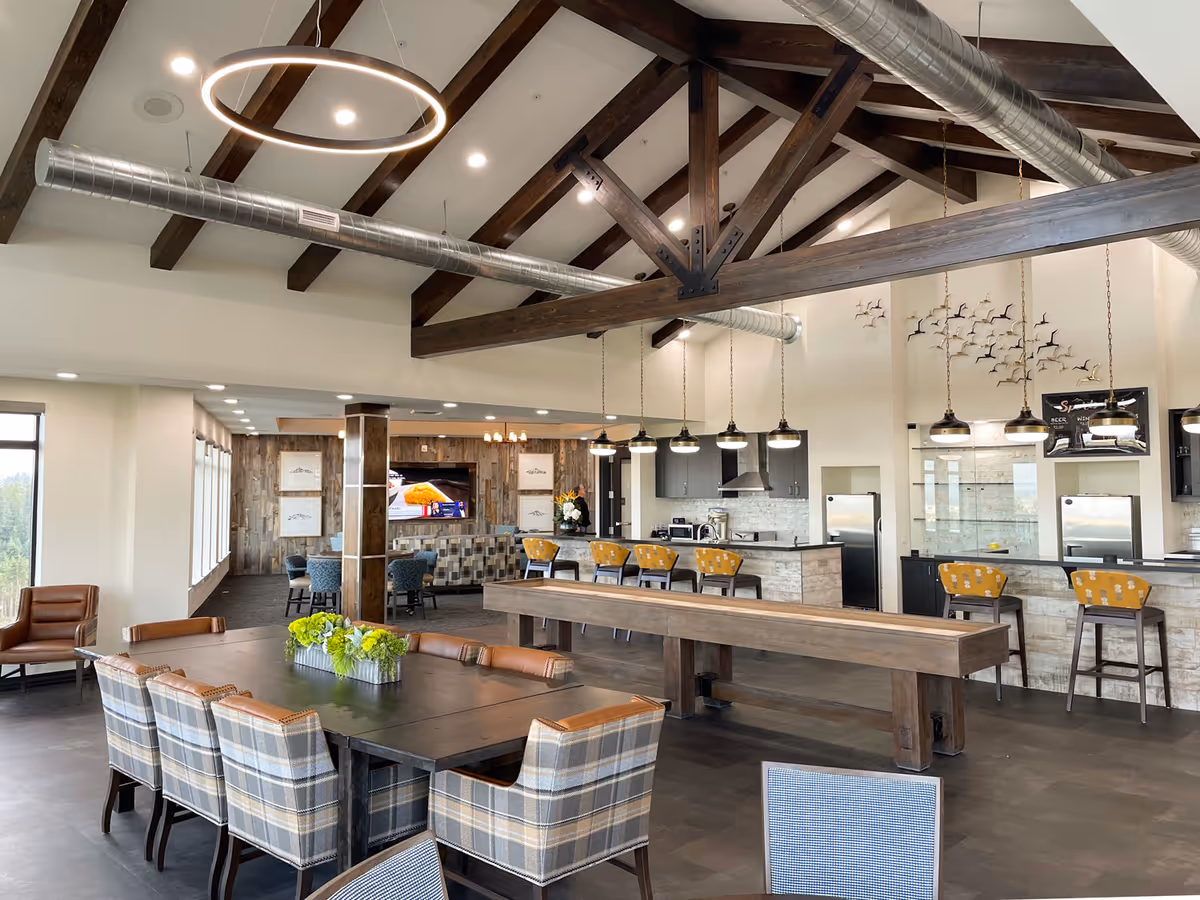 Spacious and modern common area in Boone Ridge Retirement Community featuring a large wooden table with plaid upholstered chairs, a shuffleboard table, a kitchen area with bar stools, pendant lighting, exposed wooden beams on the ceiling, and a seating area with a TV in the background.