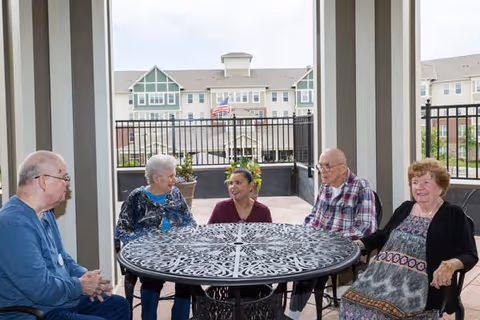 Four elderly people and one caregiver sitting around a round metal table on a covered outdoor patio with a building and American flag visible in the background.
