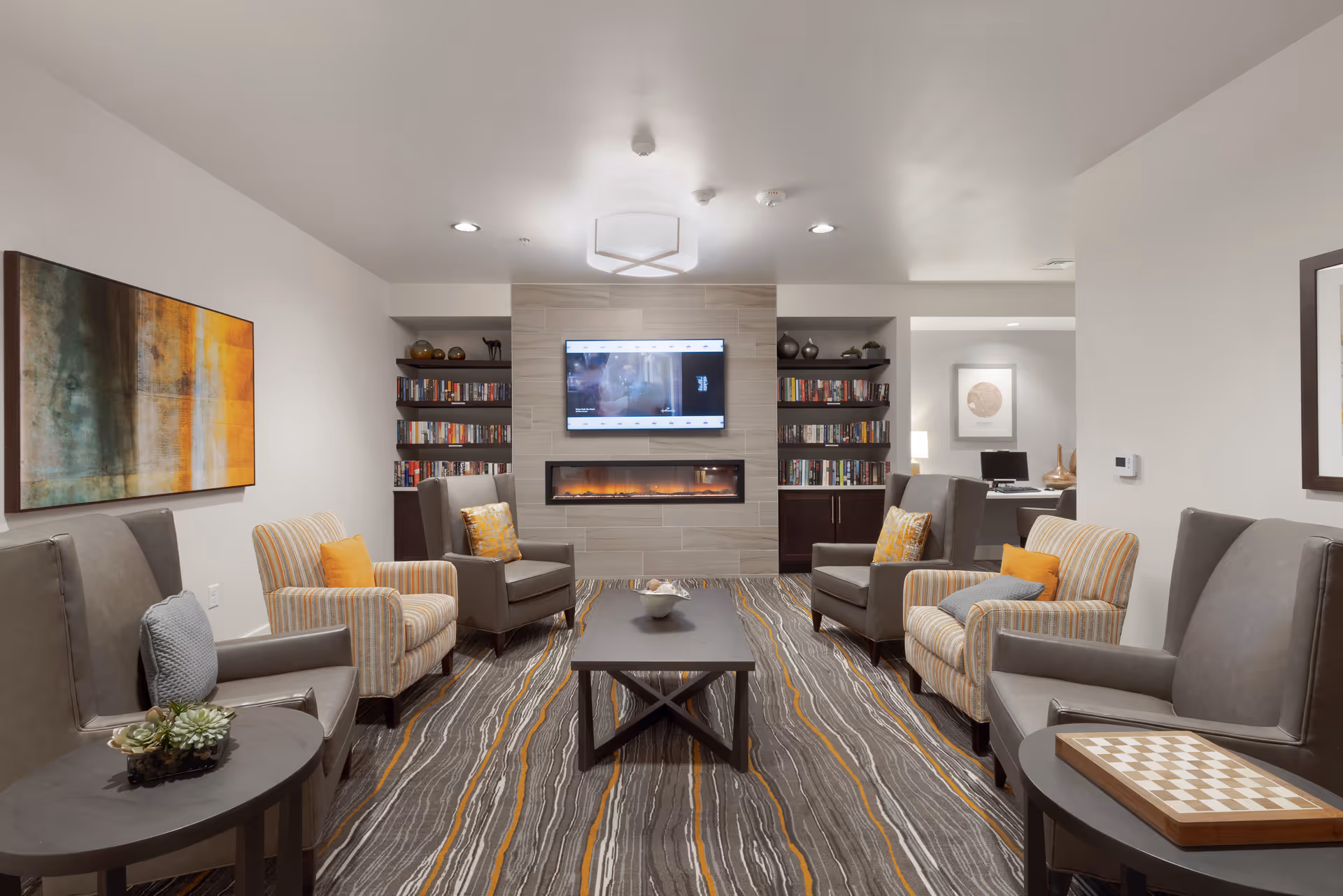 A cozy senior living common area with a modern electric fireplace and a wall-mounted TV above it. The room features a patterned carpet with gray, white, and orange stripes, several comfortable armchairs with cushions arranged around a central coffee table, built-in bookshelves filled with books on either side of the fireplace, and abstract artwork on the walls. The lighting is soft and recessed, creating a warm and inviting atmosphere.