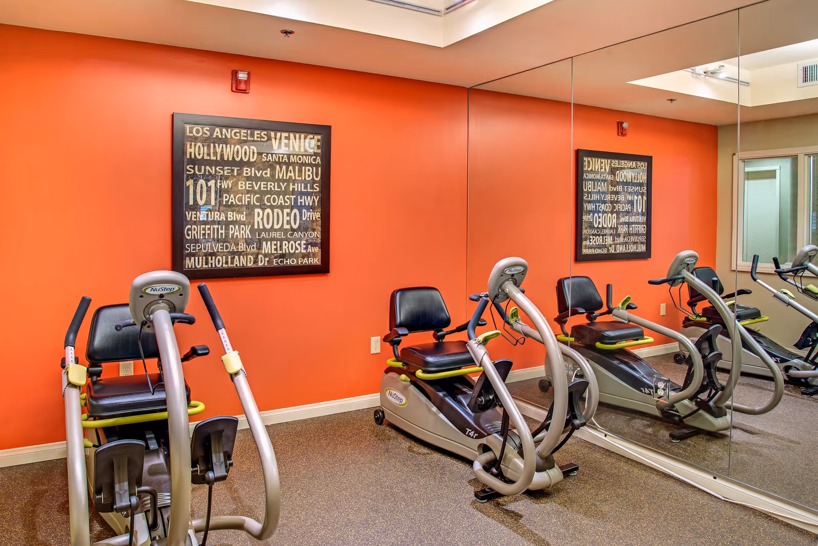 Exercise room with two NuStep recumbent cross trainers positioned in front of a large wall mirror. The walls are painted orange, and there is a framed artwork listing various Los Angeles area locations such as Venice, Hollywood, and Malibu.