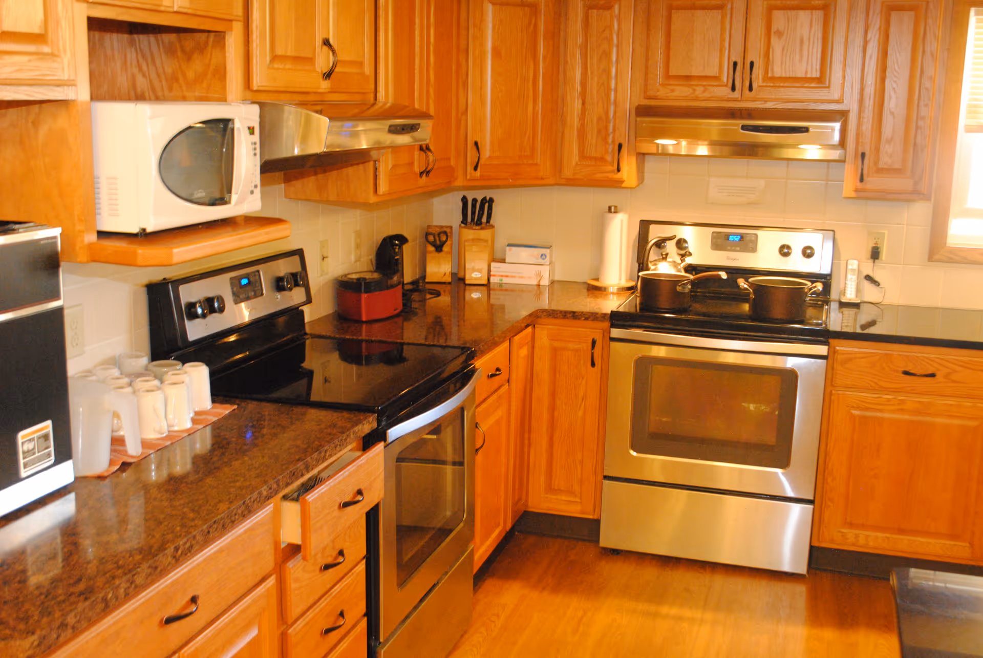 A kitchen with wooden cabinets and brown countertops. The kitchen features a stainless steel stove with two pots on it, a microwave, a coffee maker, a knife block, and several white mugs and pitchers on the counter. The floor is wooden, and there is a window with blinds partially visible on the right.