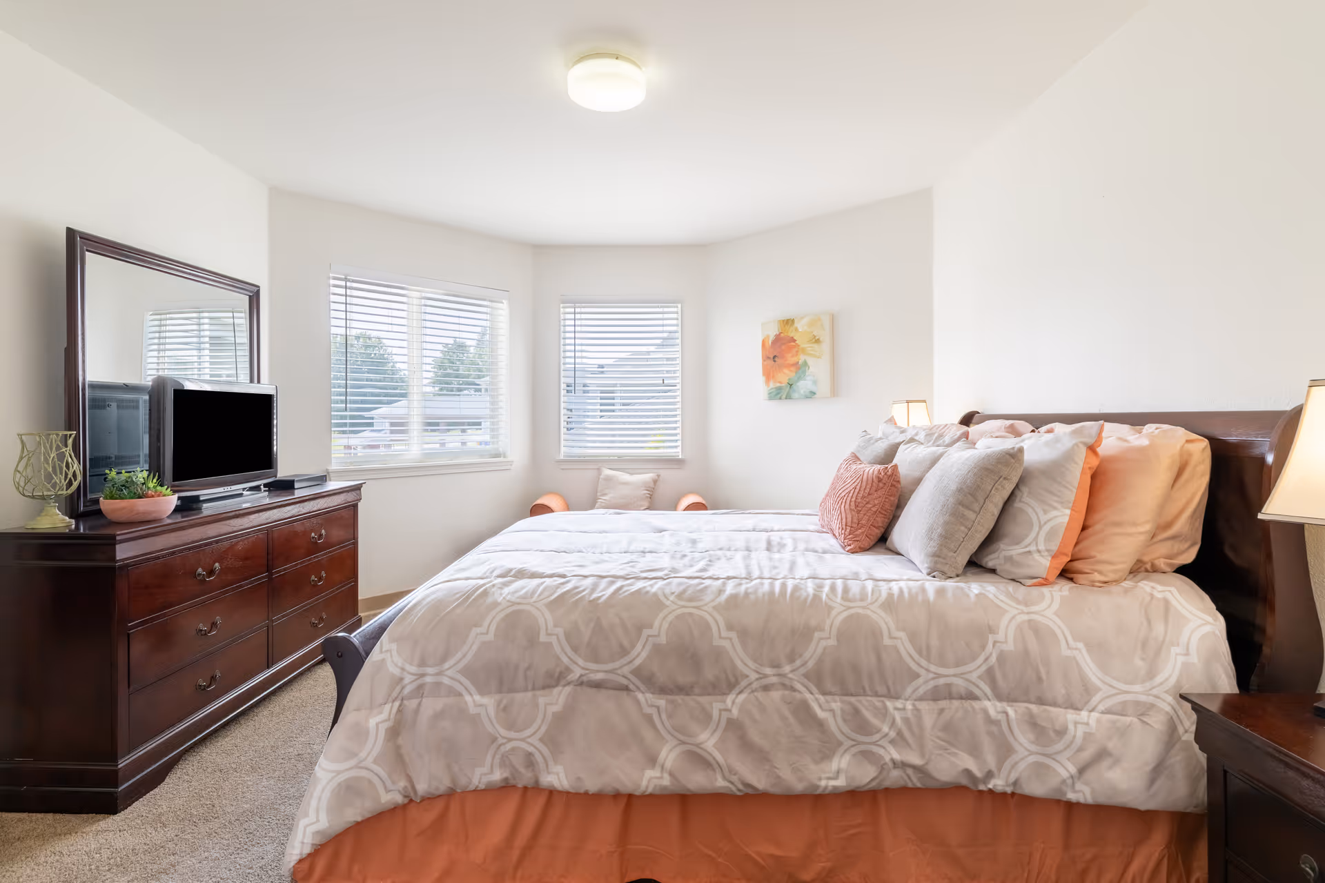 A bright bedroom with a large bed covered in a beige and white patterned comforter and multiple pillows in shades of peach and gray. There is a wooden dresser with a mirror and a small TV on top, along with decorative items. Two windows with white blinds let in natural light, and a small orange chair with a pillow is placed in the corner. A floral painting hangs on the wall above the chair.