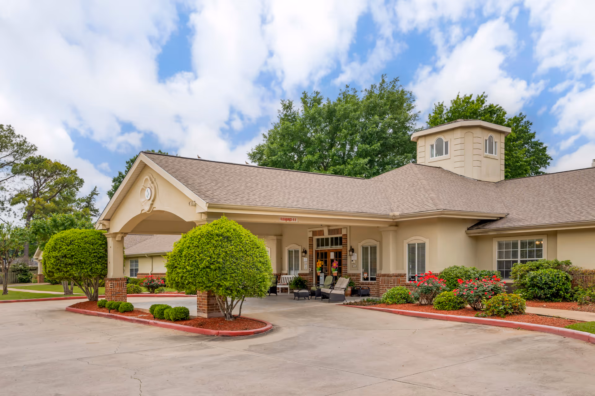 Exterior view of Brookdale Shreveport facility showing a beige building with a covered entrance, neatly trimmed bushes, red mulch landscaping, and a partly cloudy sky.