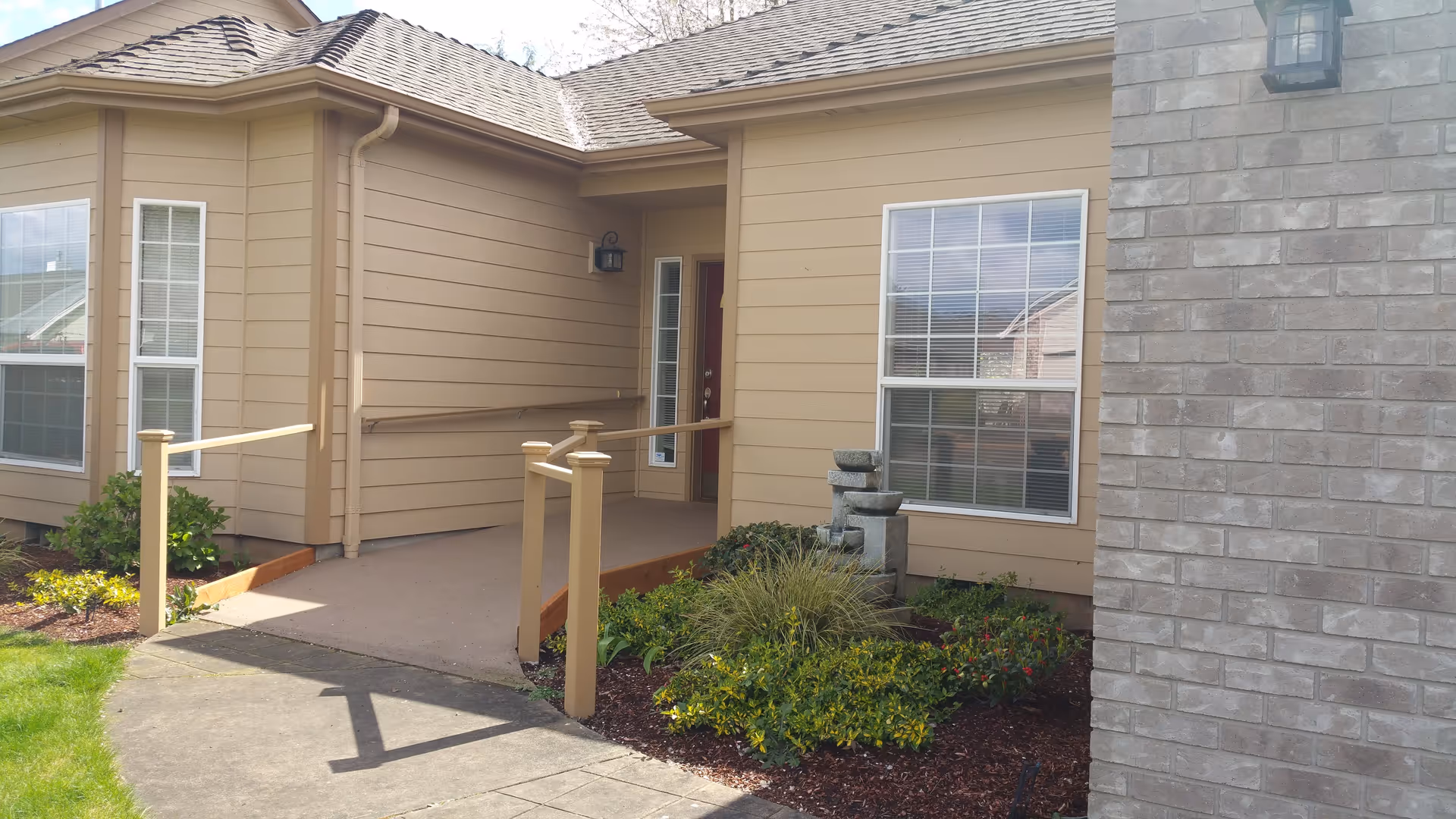 Exterior view of a beige single-story building with a sloped roof, featuring a concrete ramp with handrails leading to a door. There are two large windows with white blinds, a small garden with green plants and shrubs, and a stone water fountain near the entrance.