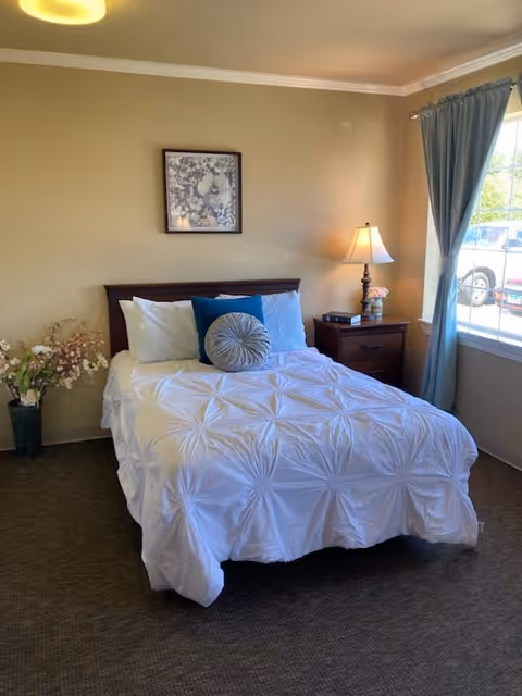 A neatly made bed with a white textured comforter and three pillows, including a round decorative pillow, in a bedroom with beige walls. There is a wooden nightstand with a lamp and a small flower arrangement next to the bed. A framed floral artwork hangs above the bed. A window with blue curtains lets in natural light, and a vase with flowers is placed on the floor near the bed.