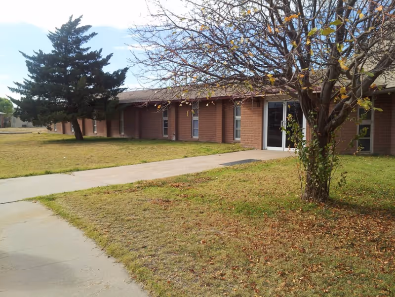 Exterior view of a single-story brick building with several windows and a glass door entrance. There is a concrete walkway leading to the entrance, a tree with sparse leaves in the foreground, and a large evergreen tree to the left. The grass around the building is patchy with some fallen leaves.