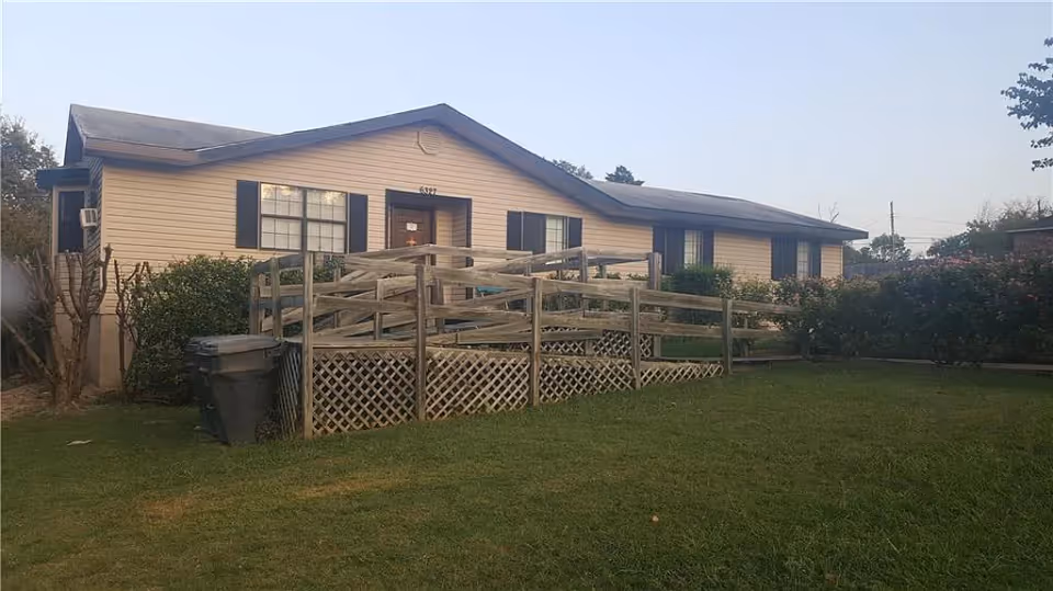 Single-story beige residential building with a wooden wheelchair ramp leading to the front door and a grassy lawn.