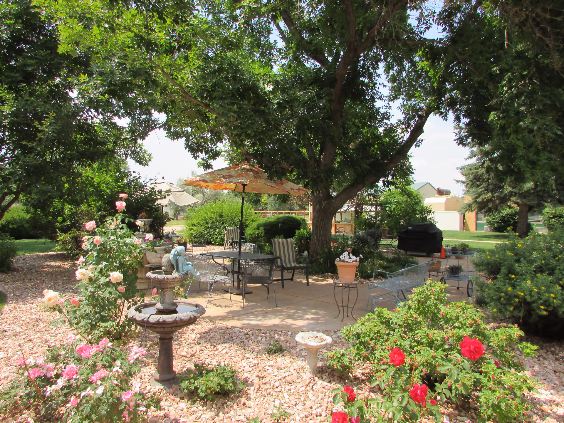 Shaded garden patio with a table, umbrella, chairs, fountains and flowering shrubs beneath large trees.