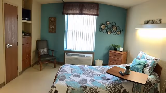 A small, cozy bedroom in a healthcare facility with a single bed covered in floral patterned bedding. There is a wooden bedside table with a plant and a small lamp, a chair with a cushion, a window with vertical blinds and a valance, and a wall decoration with circular mirrors. The walls are painted in light blue and beige tones, and a sign above the bed reads 'DREAM BIG'.
