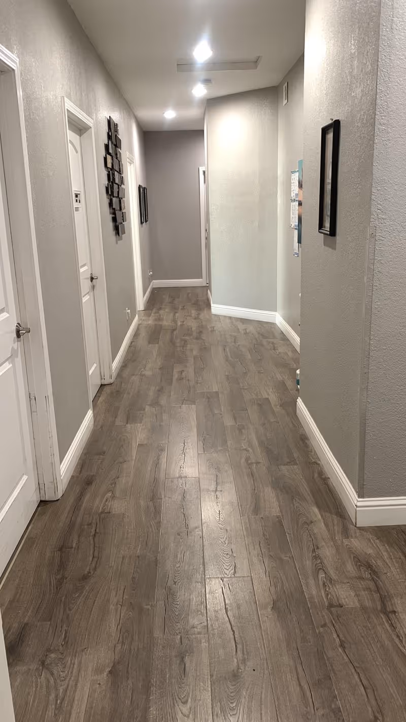 A long, narrow hallway with wood-patterned flooring and light gray walls. Several white doors line the left side of the hallway, and framed pictures or decorations hang on the walls. The ceiling has recessed lighting fixtures.