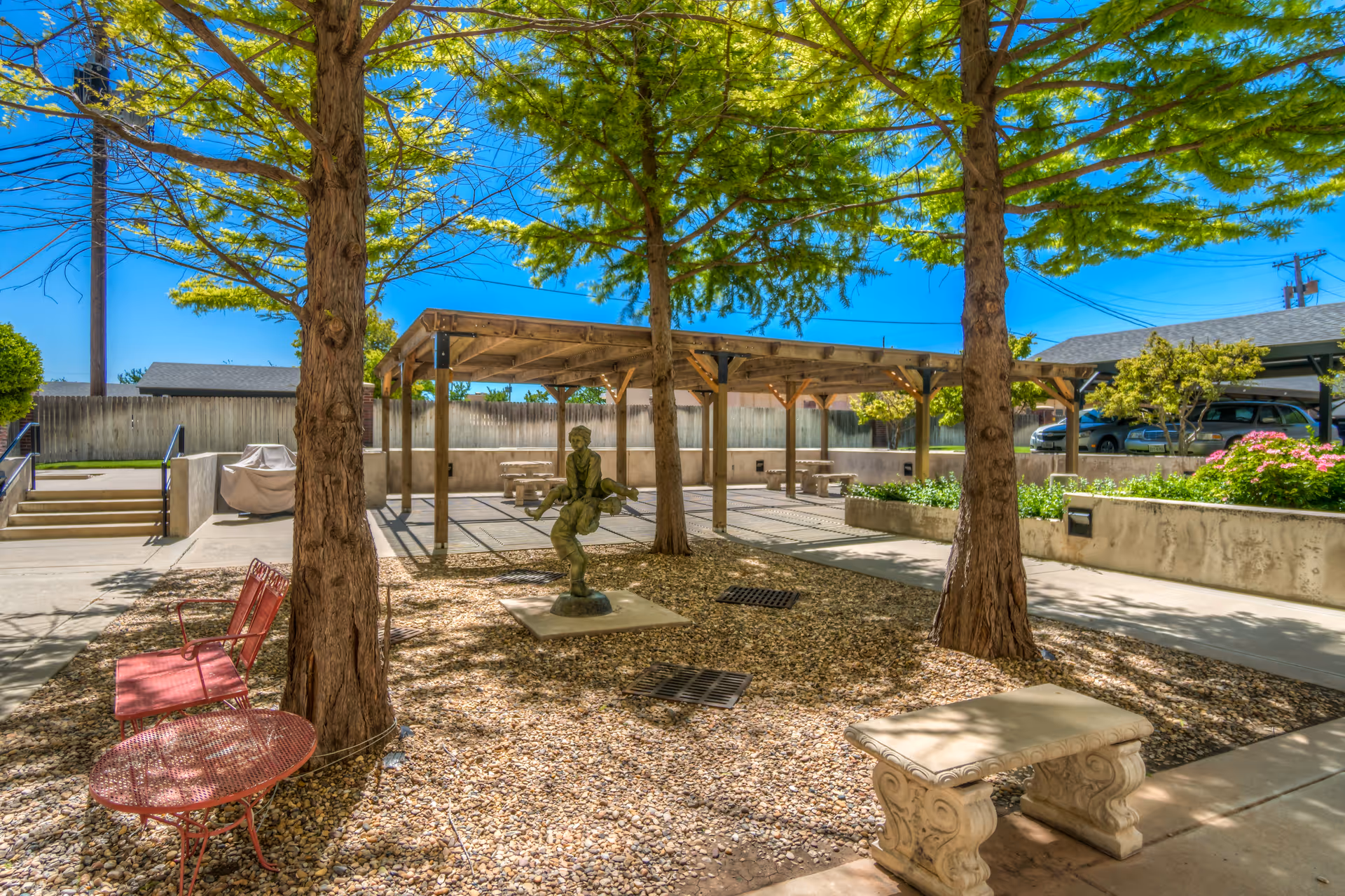 Outdoor courtyard area with trees, a statue, red metal bench and table, a stone bench, and a wooden pergola with picnic tables underneath. There are concrete walkways and a parking area with cars in the background under a clear blue sky.