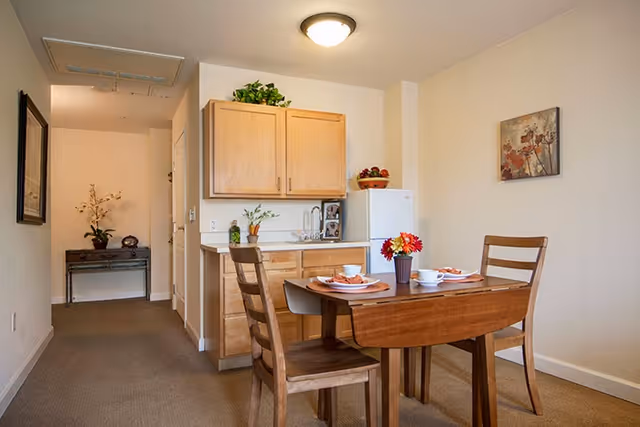 A small dining area with a wooden table set for two, featuring plates, cups, and a vase with flowers. Behind the table is a kitchenette with wooden cabinets, a small refrigerator, a coffee maker, and some decorative plants. The room has beige walls and carpeted flooring, with a framed picture on the wall and a small table with a plant in the hallway.