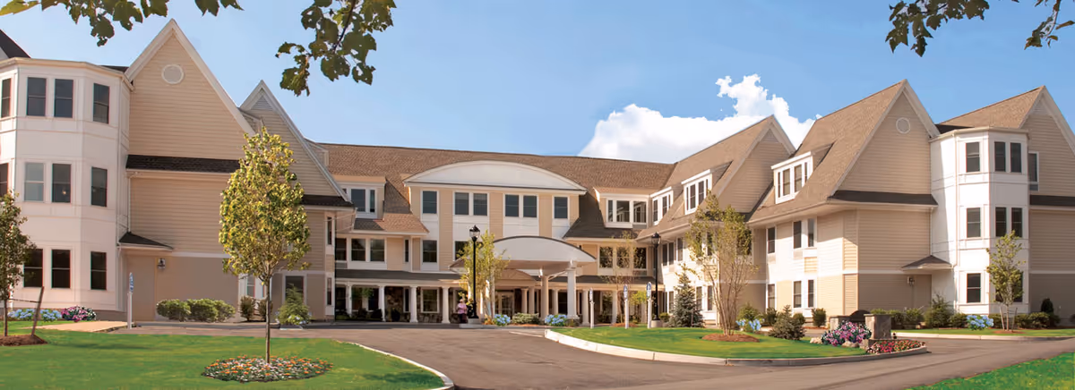 Front exterior view of a large, multi-story senior living facility building with beige siding, white trim, multiple windows, and a covered entrance. The building is surrounded by green lawns, small trees, and landscaped flower beds under a clear blue sky.