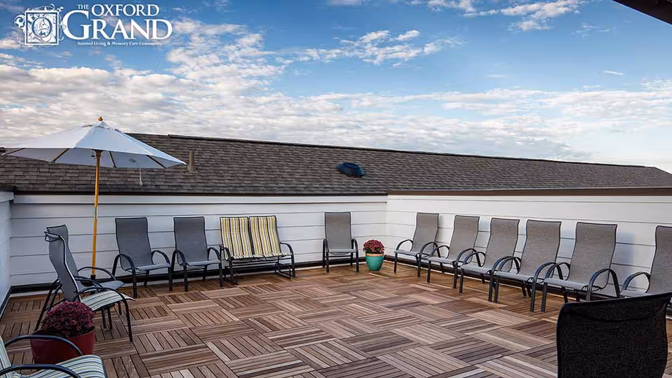Rooftop patio with a row of lounge chairs, an umbrella, potted plants and a wooden deck under a partly cloudy sky.