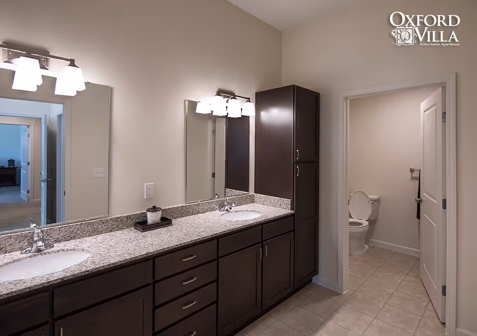 A modern bathroom with a long granite countertop featuring two sinks and dark wood cabinets underneath. Above each sink is a mirror with light fixtures. To the right, there is a separate small room with a toilet and a towel holder. The walls are painted light beige, and the floor is tiled. The logo for Oxford Villa Active Senior Apartments is visible in the upper right corner.