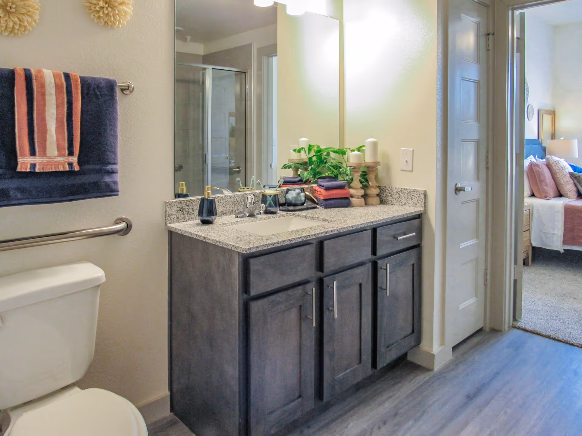 A bathroom with a toilet, a dark wood vanity with a granite countertop, a large mirror, and a towel rack holding two towels. The bathroom has a glass-enclosed shower visible in the mirror reflection. The door to an adjacent bedroom with a bed and pillows is open.