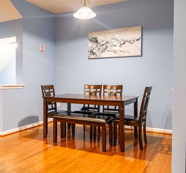 Wood dining table with four chairs and a bench against a light blue wall under a hanging light and a framed painting.