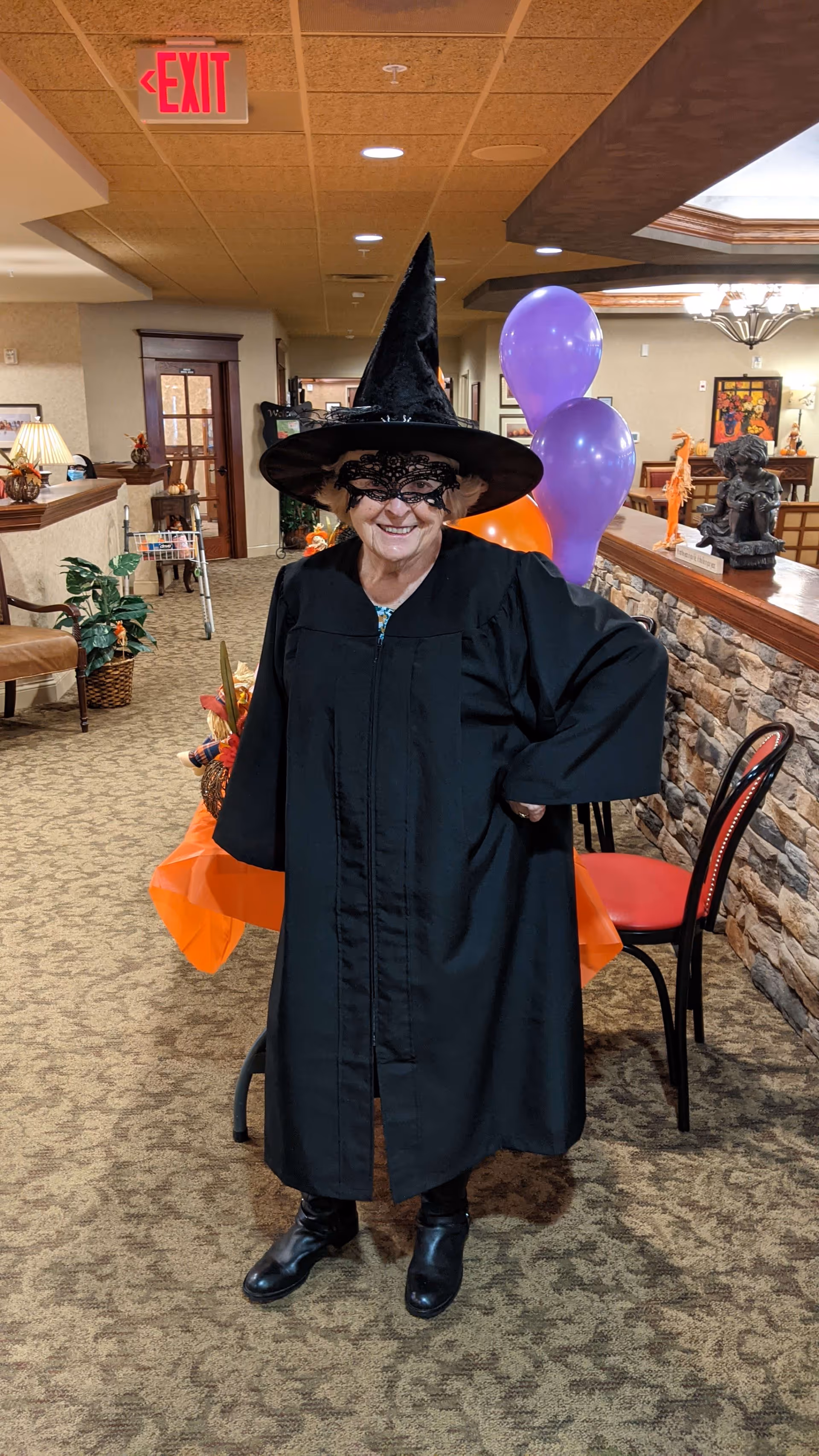 An elderly woman dressed in a black witch costume with a pointed hat and lace mask stands smiling in a decorated indoor hallway. Behind her are purple and orange balloons, a table with an orange tablecloth, and various Halloween decorations. The setting appears to be a senior living facility with carpeted floors, stone wall accents, and warm lighting.