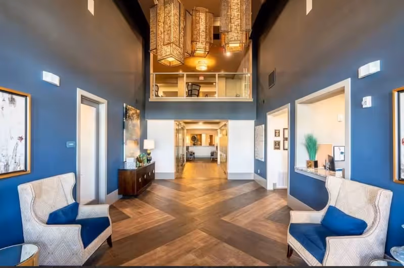Interior view of a senior living facility lobby or common area with blue walls, two beige armchairs with blue cushions, wooden flooring with a chevron pattern, a wooden console table with a lamp and decorative items, and large hanging rectangular light fixtures. There is a glass railing on the upper level and an open hallway leading further inside.