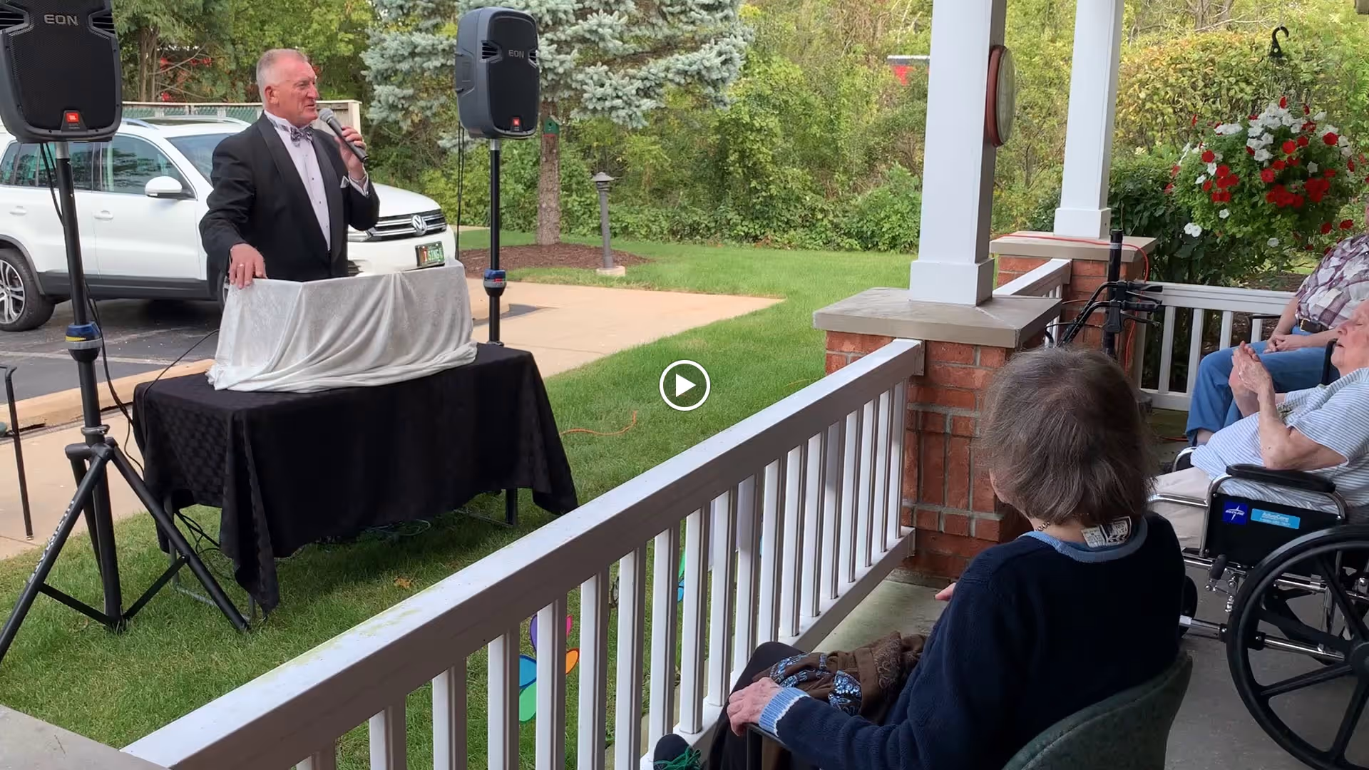 A man in a tuxedo is speaking into a microphone while standing behind a table covered with a black and white cloth on a grassy area near a porch. Several elderly people, some seated in wheelchairs, are watching him from the porch. There are speakers set up on stands, and a white vehicle is parked in the background near trees and shrubs.